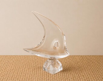 May include: A clear glass sculpture resembling a sail, mounted on a faceted base. The sail shape curves upwards, with a small hole near the center. The sculpture is displayed on a textured, neutral-toned surface.
