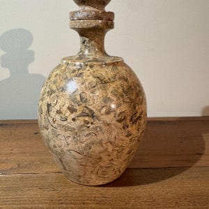 K&ouml;nnte beinhalten: Eine dekorative Steinflasche mit rundem K&ouml;rper und passendem Verschluss. Die Flasche hat ein gesprenkeltes Muster in Braun-, Beige- und Graut&ouml;nen. Der Verschluss hat eine Kugelform. Die Flasche steht auf einer Holzoberfl&auml;che.