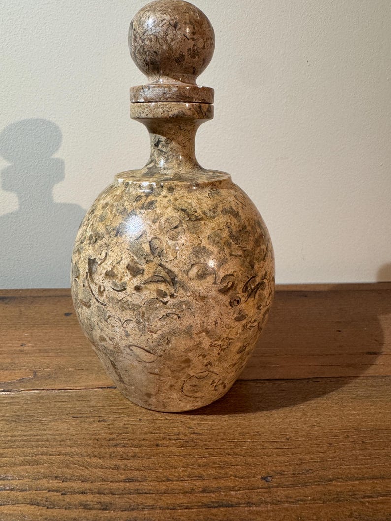 K&ouml;nnte beinhalten: Eine dekorative Steinflasche mit rundem K&ouml;rper und passendem Verschluss. Die Flasche hat ein gesprenkeltes Muster in Braun-, Beige- und Graut&ouml;nen. Der Verschluss hat eine Kugelform. Die Flasche steht auf einer Holzoberfl&auml;che.