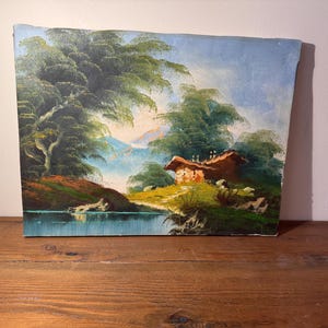 May include: Oil painting of a landscape with a small cabin, trees, and a body of water. The painting features a cabin with a brown roof, lush green trees, and a blue sky. The water reflects the surrounding scenery. The painting is on a rectangular canvas.