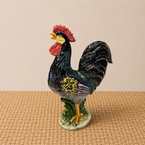 Puede incluir: Figura de gallo de cerámica con cuerpo negro, cresta roja y pico amarillo. El gallo tiene un diseño detallado con acentos de amarillo, morado y verde. Se apoya sobre una base verde con detalles de hierba, y es un objeto decorativo.