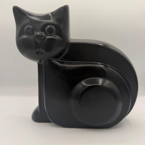 May include: A solid black ceramic cat figurine in a seated position. The cat has a smooth, glossy finish with stylized, rounded features, including large circular eyes, a small triangular nose, and a curved body shape with a prominent circular hip.