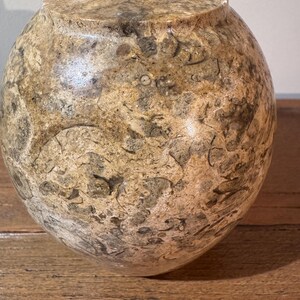 K&ouml;nnte beinhalten: Eine dekorative, runde Vase mit bauchigem K&ouml;rper und schmalem Hals. Die Vase besteht aus Stein mit einem gesprenkelten Muster aus Beige, Braun und Grau. Die Oberfl&auml;che hat eine polierte Oberfl&auml;che. Die Vase steht auf einer Holzoberfl&auml;che.