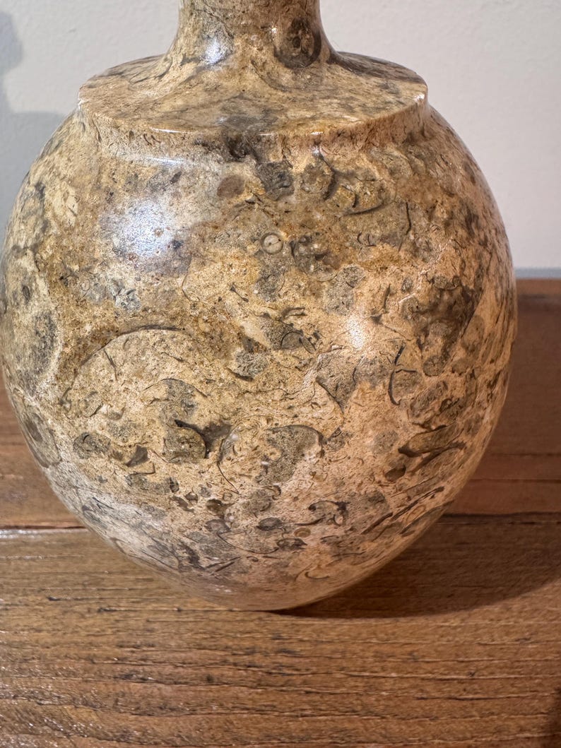 K&ouml;nnte beinhalten: Eine dekorative, runde Vase mit bauchigem K&ouml;rper und schmalem Hals. Die Vase besteht aus Stein mit einem gesprenkelten Muster aus Beige, Braun und Grau. Die Oberfl&auml;che hat eine polierte Oberfl&auml;che. Die Vase steht auf einer Holzoberfl&auml;che.