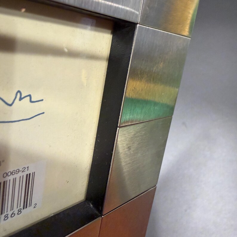 May include: A rectangular picture frame featuring a multi-toned border with brushed silver-tone metal segments and a dark brown base. The frame contains an off-white mat with a partial blue handwritten signature and a white barcode sticker labeled 0069-21.
