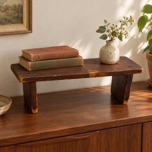 May include: A rectangular wooden riser with a dark brown, distressed finish and two sturdy legs. Two hardcover books are stacked on the left side, and a small, speckled ceramic vase containing white flowers sits on the right side.