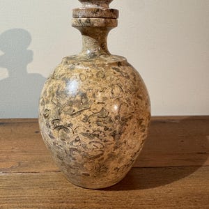 K&ouml;nnte beinhalten: Eine dekorative Steinflasche mit rundem K&ouml;rper und Verschluss. Die Flasche ist haupts&auml;chlich beige mit dunkleren braunen und grauen Adern. Der Verschluss ist eine abgerundete Kugel. Die Flasche wird wahrscheinlich zur Aufbewahrung von Fl&uuml;ssigkeiten verwendet.