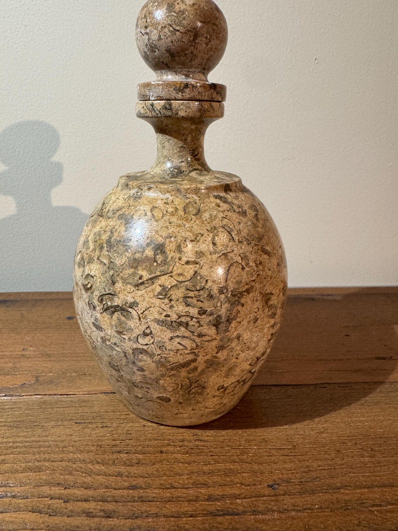 K&ouml;nnte beinhalten: Eine dekorative Steinflasche mit rundem K&ouml;rper und Verschluss. Die Flasche ist haupts&auml;chlich beige mit dunkleren braunen und grauen Adern. Der Verschluss ist eine abgerundete Kugel. Die Flasche wird wahrscheinlich zur Aufbewahrung von Fl&uuml;ssigkeiten verwendet.