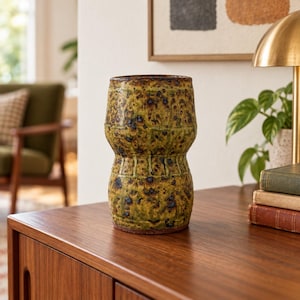 May include: A cylindrical ceramic vase with a pinched middle section, featuring a mottled olive green and brown glaze with dark speckles and a textured surface. The vase has a wide opening at the top and a flat base, displaying a rustic finish.