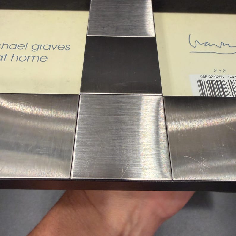 May include: A rectangular picture frame with a brushed metal finish and a black border. The cream-colored insert features the text Michael Graves at home and a signature. A barcode label indicates a 3 inch by 3 inch photo size for the frame.