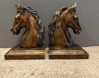 May include: A pair of bookends featuring sculpted horse heads in a metallic bronze finish. Each horse head is mounted on a rectangular base, showcasing detailed manes and facial features with a weathered, antique-style patina.