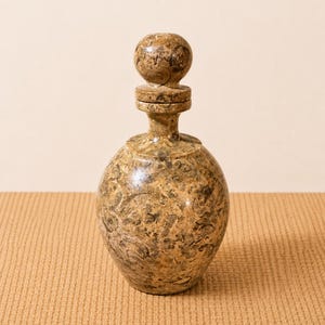 K&ouml;nnte beinhalten: Eine dekorative, runde Flasche mit passendem Verschluss. Die Flasche und der Verschluss bestehen aus einem gesprenkelten, hellbraunen Material mit dunkleren braunen und schwarzen Markierungen. Die Flasche hat einen bauchigen K&ouml;rper und einen schlanken Hals.