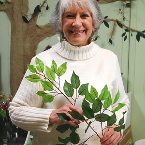 May include: A woman wearing a white turtleneck sweater holds a branch of artificial green leaves. The leaves are arranged in a fan shape, with the branch extending to the right.