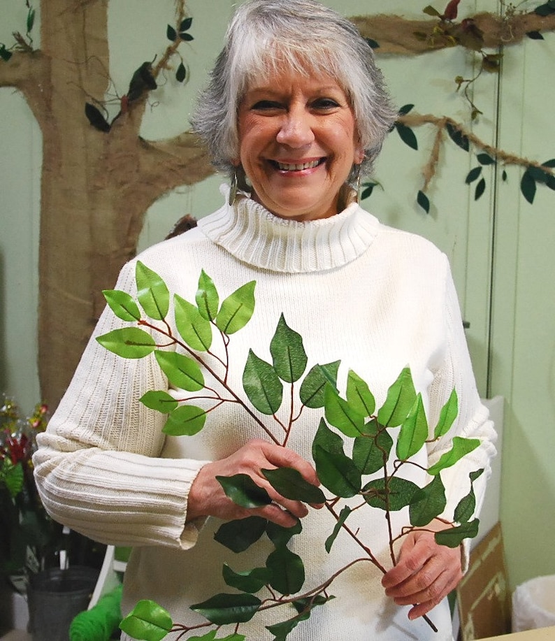 May include: A woman wearing a white turtleneck sweater holds a branch of artificial green leaves. The leaves are arranged in a fan shape, with the branch extending to the right.