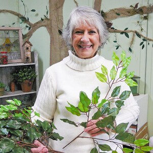 May include: A woman holds two sprigs of artificial greenery with green leaves. The sprigs are similar in color and shape. The woman is wearing a white turtleneck sweater and smiling.