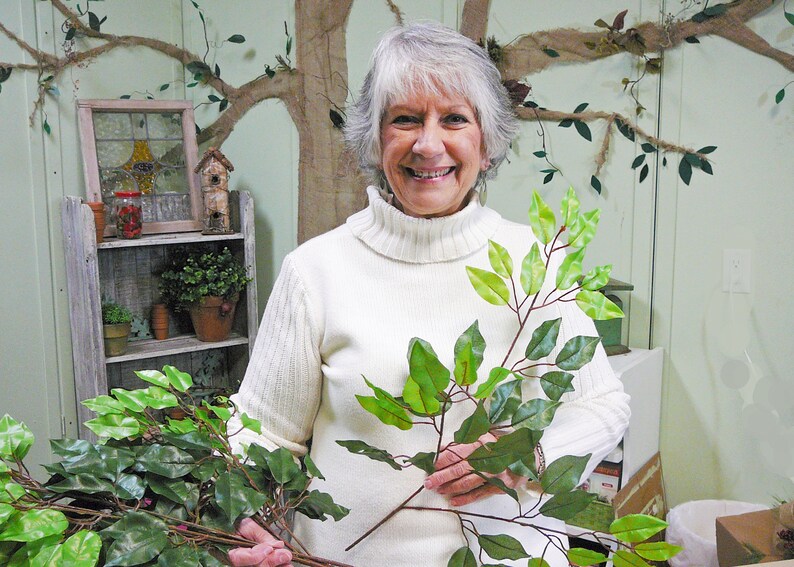 May include: A woman holds two sprigs of artificial greenery with green leaves. The sprigs are similar in color and shape. The woman is wearing a white turtleneck sweater and smiling.