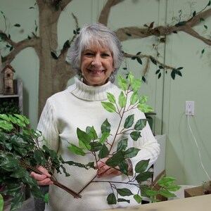 May include: A woman holds two sprigs of artificial greenery with dark green leaves. The sprigs are similar in size and shape. The woman is wearing a white turtleneck sweater and is smiling at the camera.