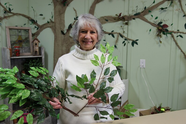 May include: A woman holds two sprigs of artificial greenery with dark green leaves. The sprigs are similar in size and shape. The woman is wearing a white turtleneck sweater and is smiling at the camera.