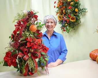 May include: A festive floral arrangement featuring red poinsettias, apples, and orange-colored fruit, accented with green foliage and red ribbon. The arrangement is displayed in a dark brown container, creating a holiday-themed centerpiece.