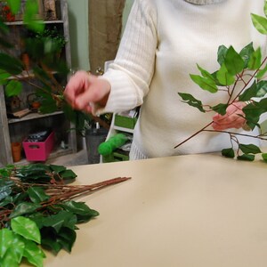 May include: A person is holding two bunches of artificial green leaves with brown stems. The leaves are arranged in a fan shape. The person is wearing a white sweater.