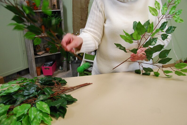 May include: A person is holding two bunches of artificial green leaves with brown stems. The leaves are arranged in a fan shape. The person is wearing a white sweater.