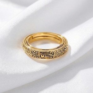 May include: A gold-colored ring with an intricate, engraved design. The ring features a series of repeating patterns and a small, raised circular detail. The ring is set against a white, textured background.