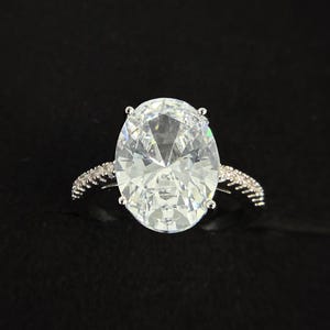 May include: A silver ring with a large, oval-cut, clear gemstone as the centerpiece. The band is adorned with smaller, clear stones. The ring is set against a black background, highlighting its sparkle and design.