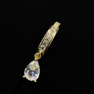 May include: A gold earring with a clear, teardrop-shaped gemstone pendant. The hoop design is adorned with small, clear stones. The pendant hangs from a delicate gold chain, creating a sparkling effect. The earring is set against a black background.