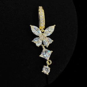 May include: A gold-toned earring with a butterfly design and clear, sparkling stones. The earring features a hoop at the top, a butterfly-shaped centerpiece, a square stone, and a round stone dangling below. The background is black.