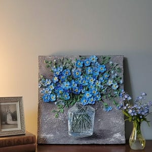 May include: A textured painting of blue flowers in a clear vase. The artwork is on a grey background, and a small vase of flowers sits to the right. A framed photo and books are on a wooden surface.