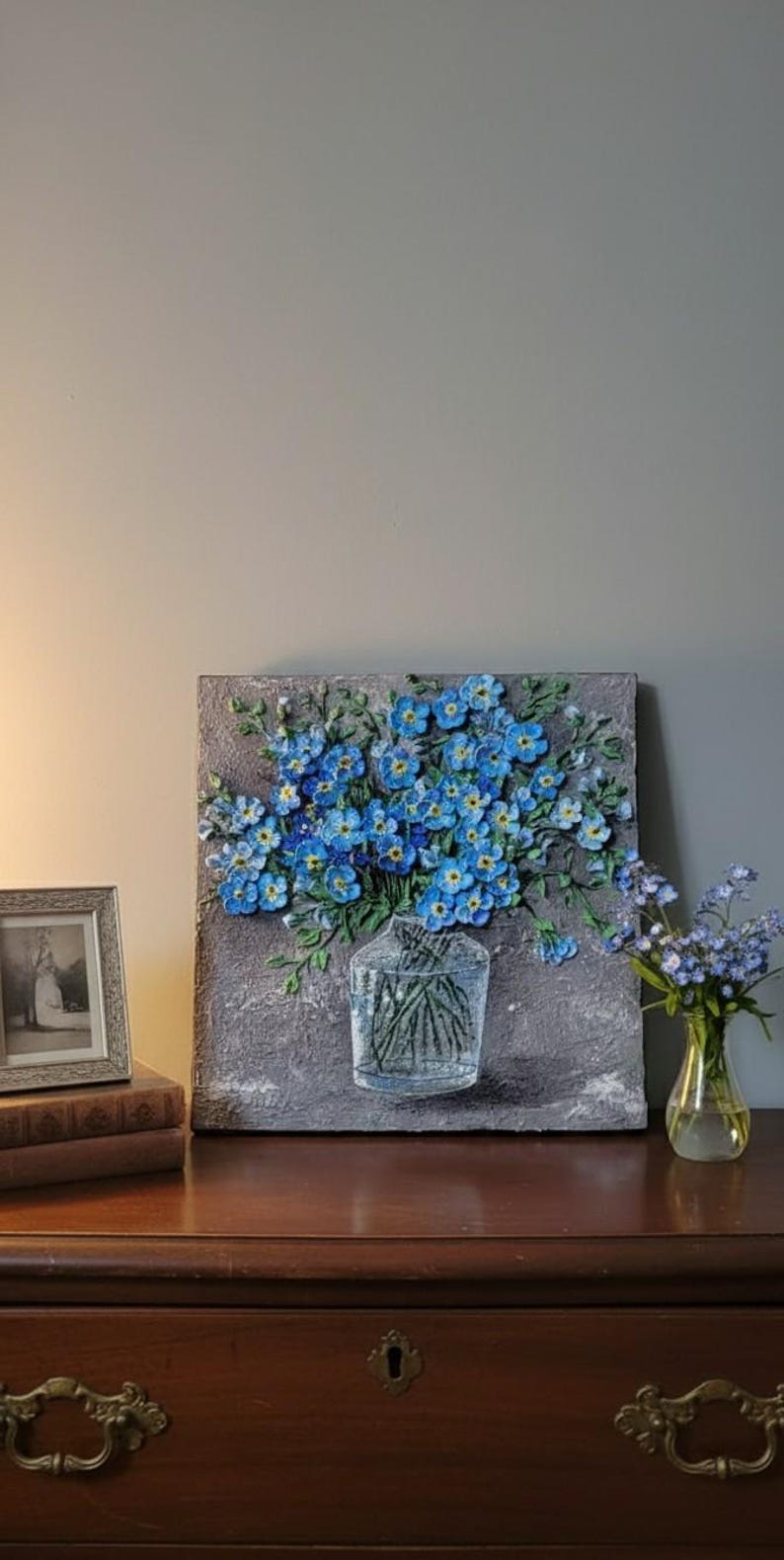 May include: A textured painting of blue flowers in a clear vase. The artwork is on a grey background, and a small vase of flowers sits to the right. A framed photo and books are on a wooden surface.