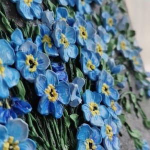 May include: A 3D relief sculpture of blue forget-me-not flowers with yellow centres and green stems. The flowers are clustered, creating a textured effect against a grey background.