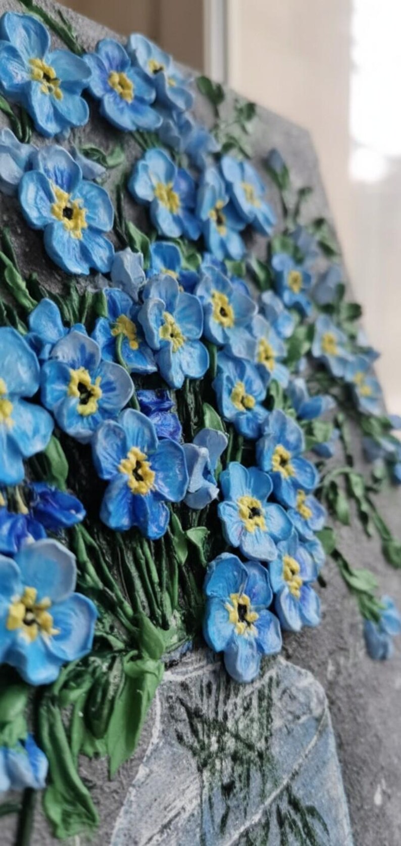 May include: A 3D relief sculpture of blue forget-me-not flowers with yellow centres and green stems. The flowers are clustered, creating a textured effect against a grey background.