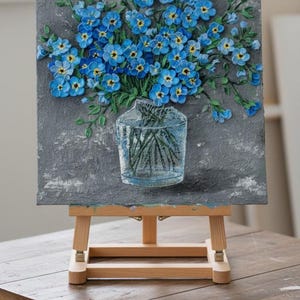May include: A painting of blue forget-me-nots in a clear glass vase. The flowers have yellow centres and green stems, set against a textured grey background. The artwork is displayed on a wooden easel.