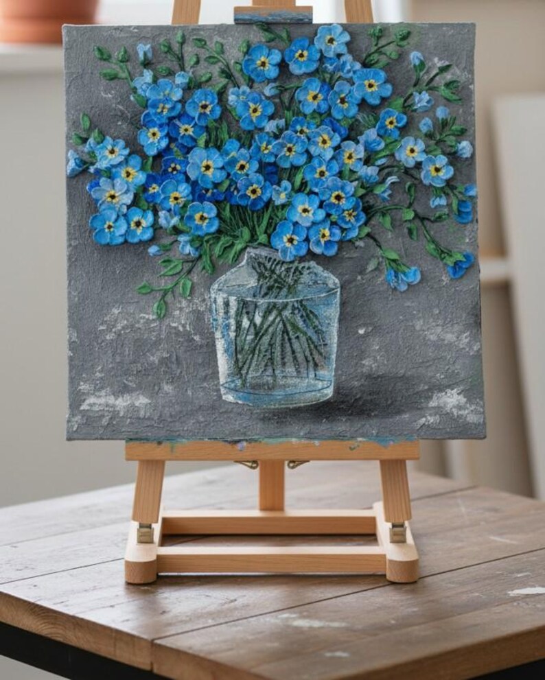 May include: A painting of blue forget-me-nots in a clear glass vase. The flowers have yellow centres and green stems, set against a textured grey background. The artwork is displayed on a wooden easel.