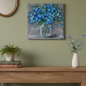 May include: A textured painting of blue flowers in a clear glass vase. The artwork is mounted on a grey background and hangs on a green wall. A small white vase with flowers sits on a wooden table.