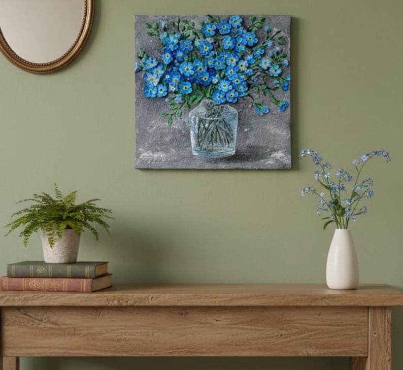 May include: A textured painting of blue flowers in a clear glass vase. The artwork is mounted on a grey background and hangs on a green wall. A small white vase with flowers sits on a wooden table.