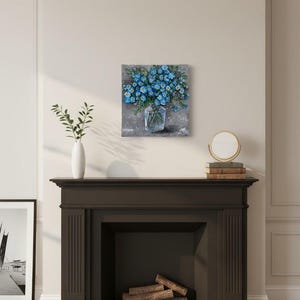 May include: A square painting shows a bouquet of blue flowers in a clear glass vase. The artwork is set against a textured grey background. The painting is displayed above a dark brown fireplace.