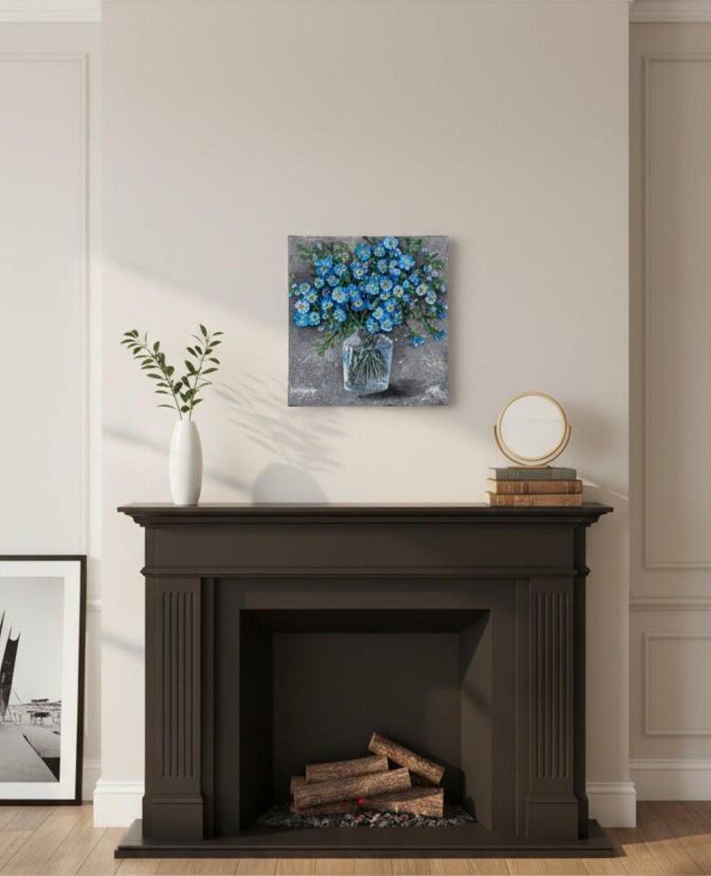 May include: A square painting shows a bouquet of blue flowers in a clear glass vase. The artwork is set against a textured grey background. The painting is displayed above a dark brown fireplace.
