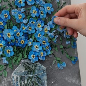 May include: A textured painting of blue forget-me-nots with yellow centres in a clear vase. The artwork is on a grey background and is displayed on a wooden easel. The flowers are in full bloom, with green leaves and stems.