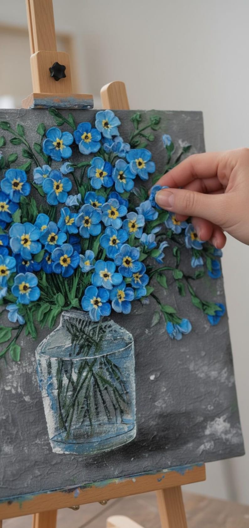 May include: A textured painting of blue forget-me-nots with yellow centres in a clear vase. The artwork is on a grey background and is displayed on a wooden easel. The flowers are in full bloom, with green leaves and stems.