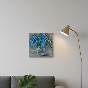 May include: Square canvas art depicting blue flowers in a clear glass vase. The painting has a textured, impressionistic style with a grey background. The artwork is displayed on a white wall above a grey sofa.