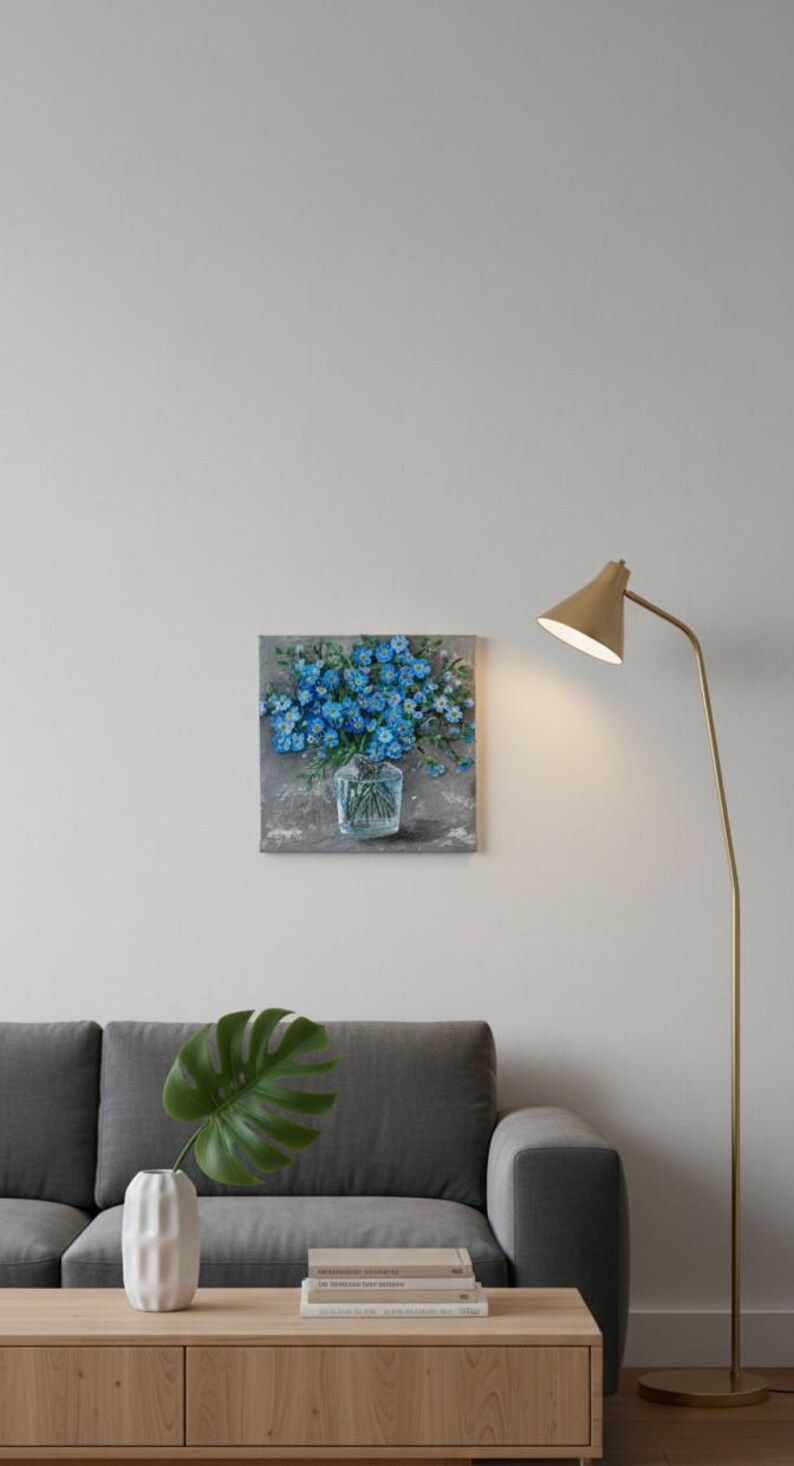 May include: Square canvas art depicting blue flowers in a clear glass vase. The painting has a textured, impressionistic style with a grey background. The artwork is displayed on a white wall above a grey sofa.