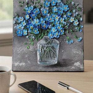 May include: A textured square artwork with a 3D relief of blue forget-me-not flowers in a clear glass vase. The flowers have yellow centres and green stems, set against a grey background. A coffee mug, phone, and pen are on the wooden surface.