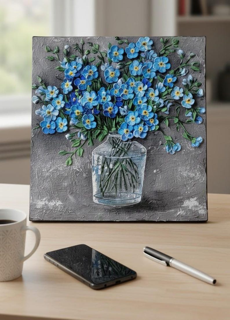 May include: A textured square artwork with a 3D relief of blue forget-me-not flowers in a clear glass vase. The flowers have yellow centres and green stems, set against a grey background. A coffee mug, phone, and pen are on the wooden surface.