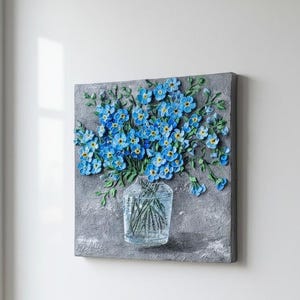 May include: A square canvas artwork with a textured grey background. A clear glass vase holds green stems and a bouquet of blue flowers with yellow centres. The artwork is mounted on a white wall.