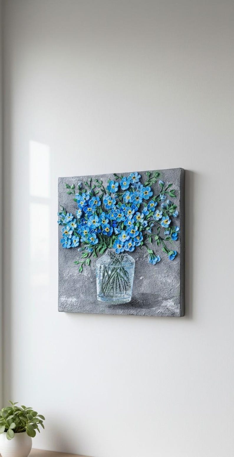 May include: A square canvas artwork with a textured grey background. A clear glass vase holds green stems and a bouquet of blue flowers with yellow centres. The artwork is mounted on a white wall.