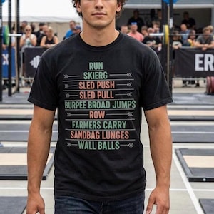 Può includere: T-shirt girocollo nero con un elenco di esercizi di allenamento stampati in bianco, verde e rosso. Gli esercizi includono Run, Skierg, Sled Push, Sled Pull, Burpee Broad Jumps, Row, Farmers Carry, Sandbag Lunges e Wall Balls.