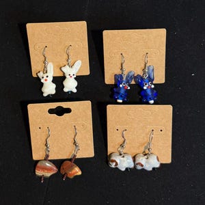 May include: Four pairs of earrings displayed on brown cards. The earrings feature various designs, including white and blue bunny figures, gemstone beads, and white elephant figures. The earrings are on silver hooks.
