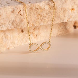 May include: A gold necklace with an infinity symbol pendant, set with small, clear gemstones. The chain is fine, and the pendant is centrally placed. The necklace is shown against a neutral, textured backdrop.